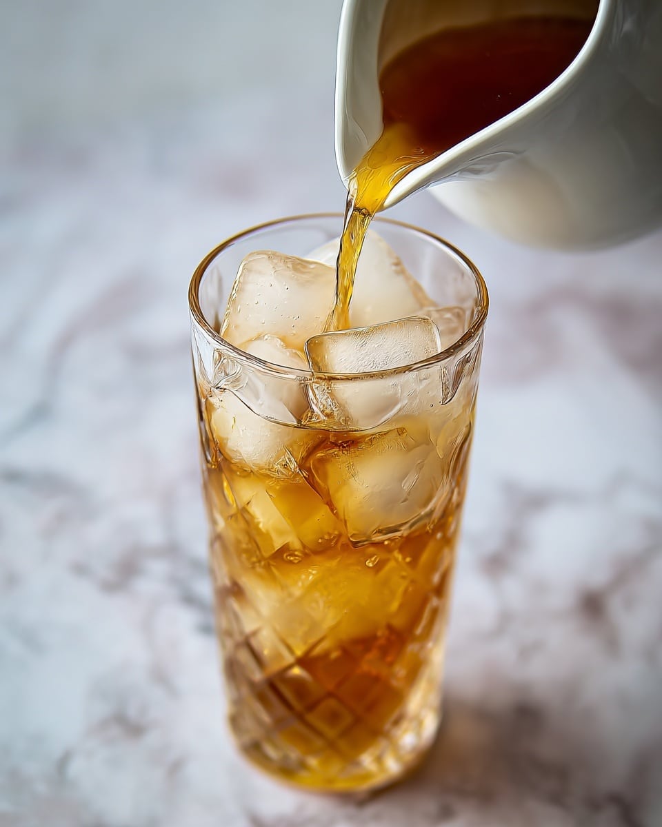 A tall clear glass filled with large ice cubes that are slightly frosty, the top layer showing five ice cubes closely packed. A golden-brown liquid is being poured into the glass from a white ceramic pitcher, with the liquid flowing smoothly and some bubbles forming near the ice. The glass shows a textured pattern that catches light, giving it a sparkling look. The background is a white marbled surface with soft blurred details. photo taken with an iphone --ar 4:5 --v 7