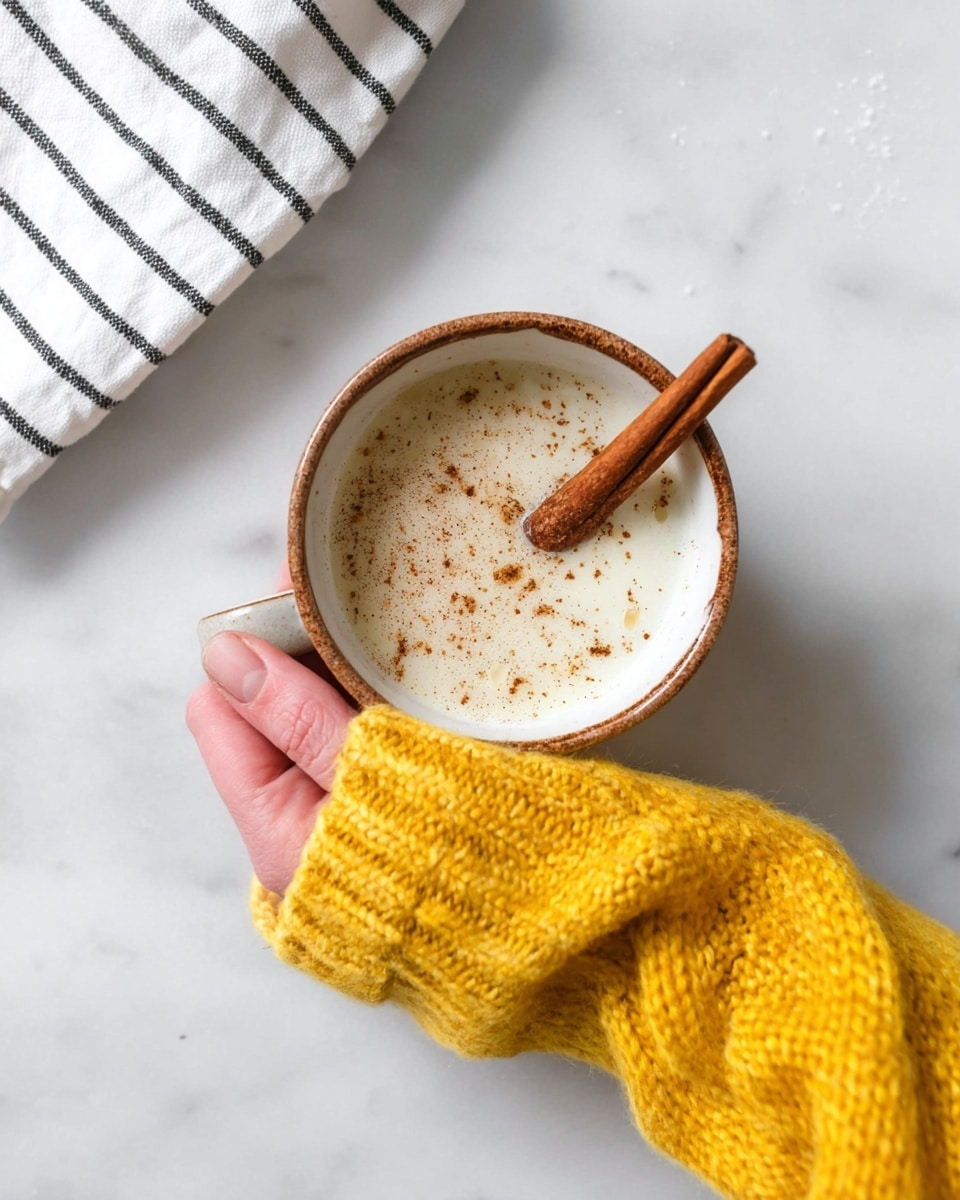 A white cup with a brown rim is held by a woman's hand wearing a bright yellow knitted sweater. The cup is filled with a light frothy drink, sprinkled with small brown specks, likely cinnamon or nutmeg, with a single cinnamon stick resting on the surface. The scene is set on a white marbled surface with a black and white striped cloth partially visible in the top left corner. photo taken with an iphone --ar 4:5 --v 7