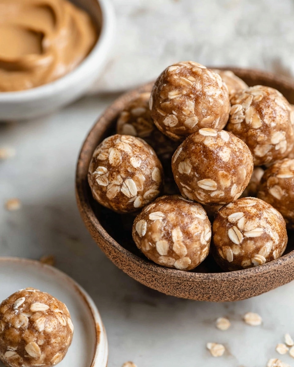 A close-up view of a bowl filled with round energy balls that have a light brown color and are studded with visible whole oats on the surface. Each ball is coated with a fine dusting of cinnamon or similar spice, giving a slightly textured look. The bowl is brown and rustic, contrasting with the white marbled texture surface underneath. In the background, there is a white bowl partially visible with a scoop of light brown peanut butter, and another white plate with one energy ball on it, positioned at the lower right corner of the image. Photo taken with an iphone --ar 4:5 --v 7