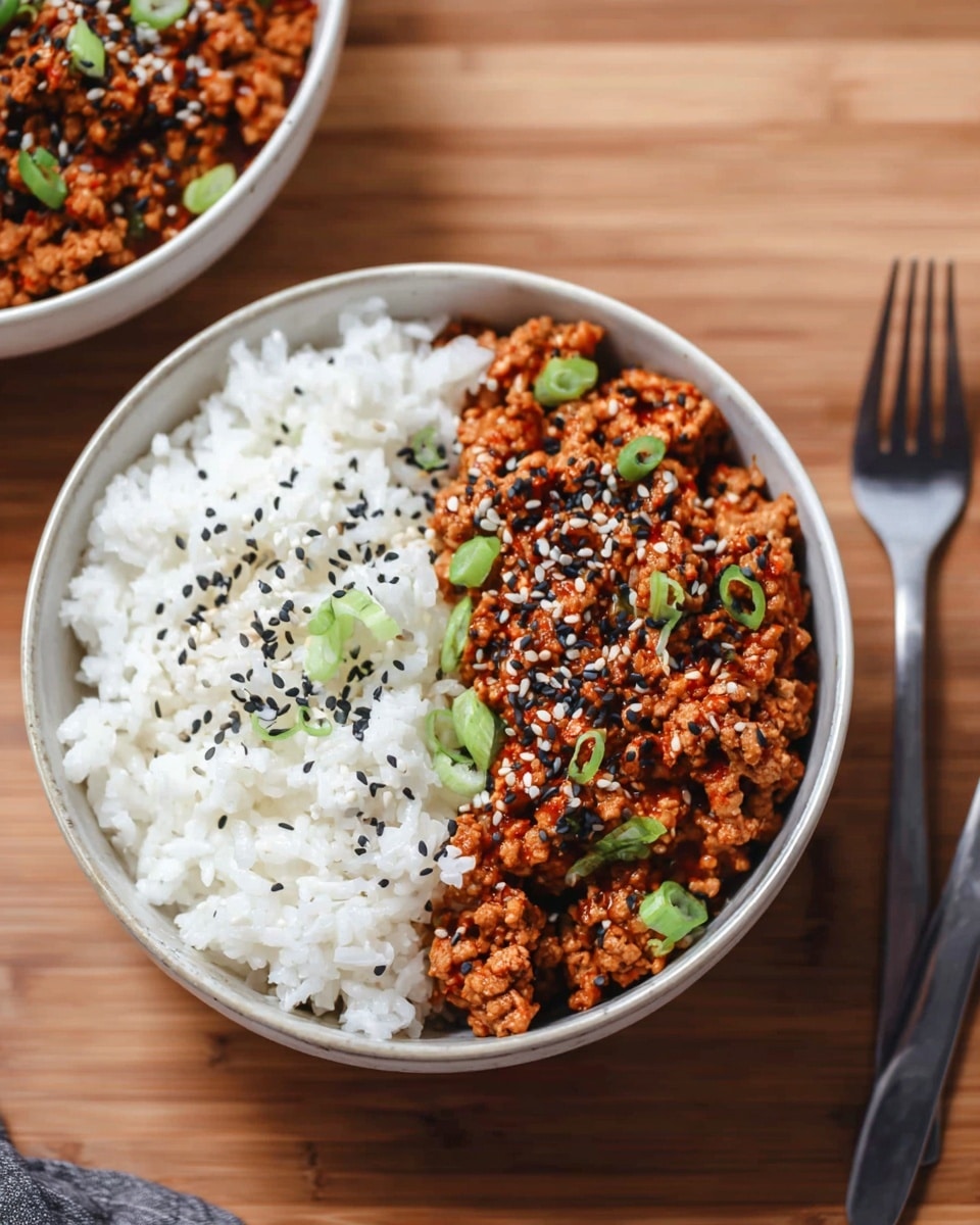 A white bowl shows two main layers: on one side, there is a layer of cooked white rice with soft, fluffy grains, and on the other side, there is a layer of cooked ground meat in a rich, reddish-orange sauce, speckled with black spots and garnished with chopped green onions and white and black sesame seeds on top. The bowl is placed on a wooden surface next to another similar bowl with the same food and a fork with a dark handle lies nearby. photo taken with an iphone --ar 4:5 --v 7