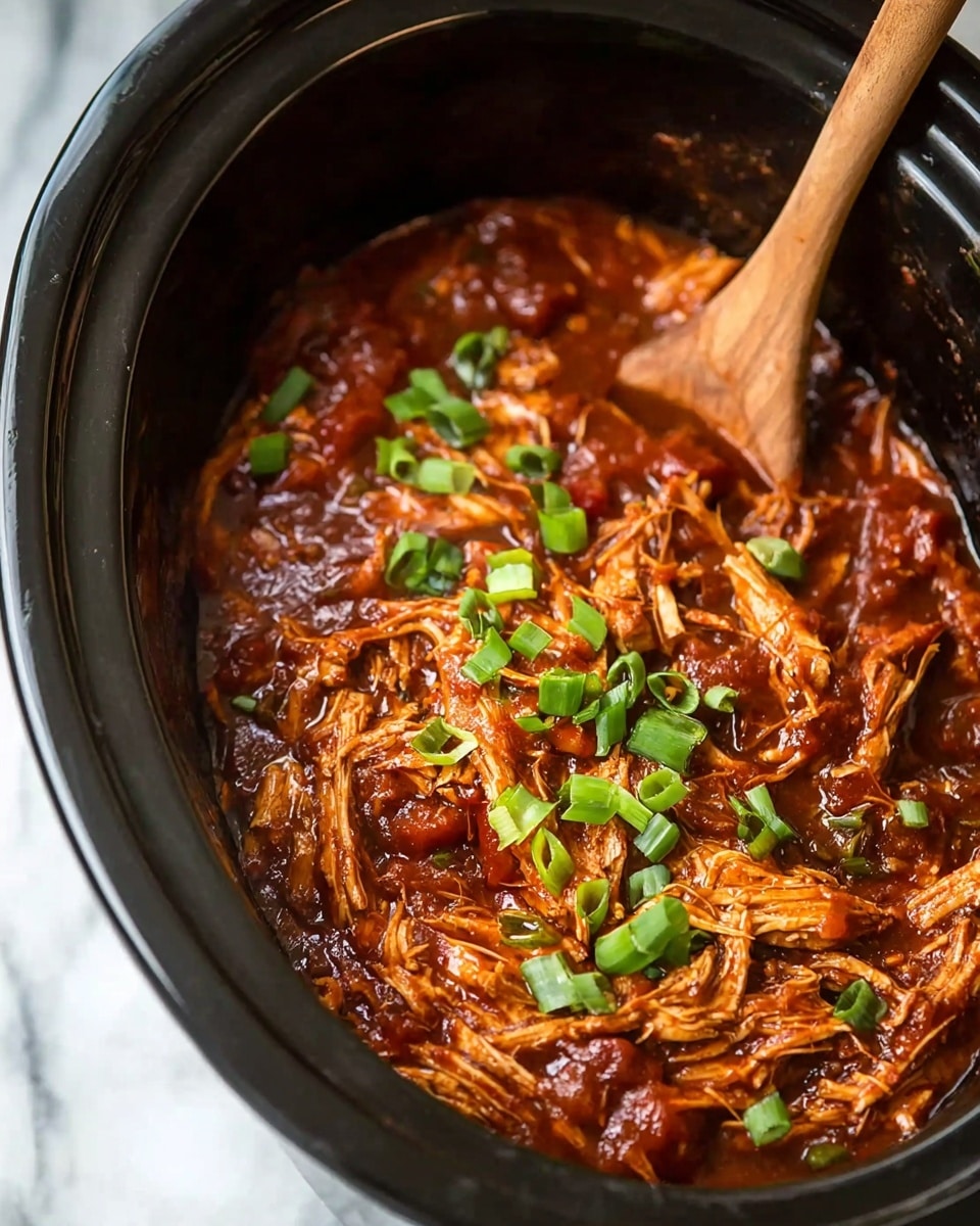 A close-up view inside a black slow cooker filled with shredded, tender chicken in a thick, rich, dark reddish-brown sauce. The sauce looks smooth and glossy, coating the chicken pieces well, with chunks of tomato or similar ingredients mixed in. Bright green, chopped spring onions are sprinkled on top, adding a fresh contrast to the dish. A wooden spoon is partially visible in the upper right corner, stirring the mixture. The slow cooker sits on a white marbled surface. photo taken with an iphone --ar 4:5 --v 7