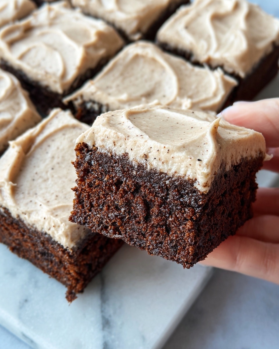A close-up view of a square chocolate cake cut into eight pieces on a white marbled surface. Each piece has two layers: a thick, dark brown, moist cake layer at the bottom, and a creamy light beige frosting layer on top with small darker specks, spread smoothly but with visible swirls. A woman's hand holding one piece of the cake shows the soft texture and the thickness of both layers clearly. Photo taken with an iphone --ar 4:5 --v 7