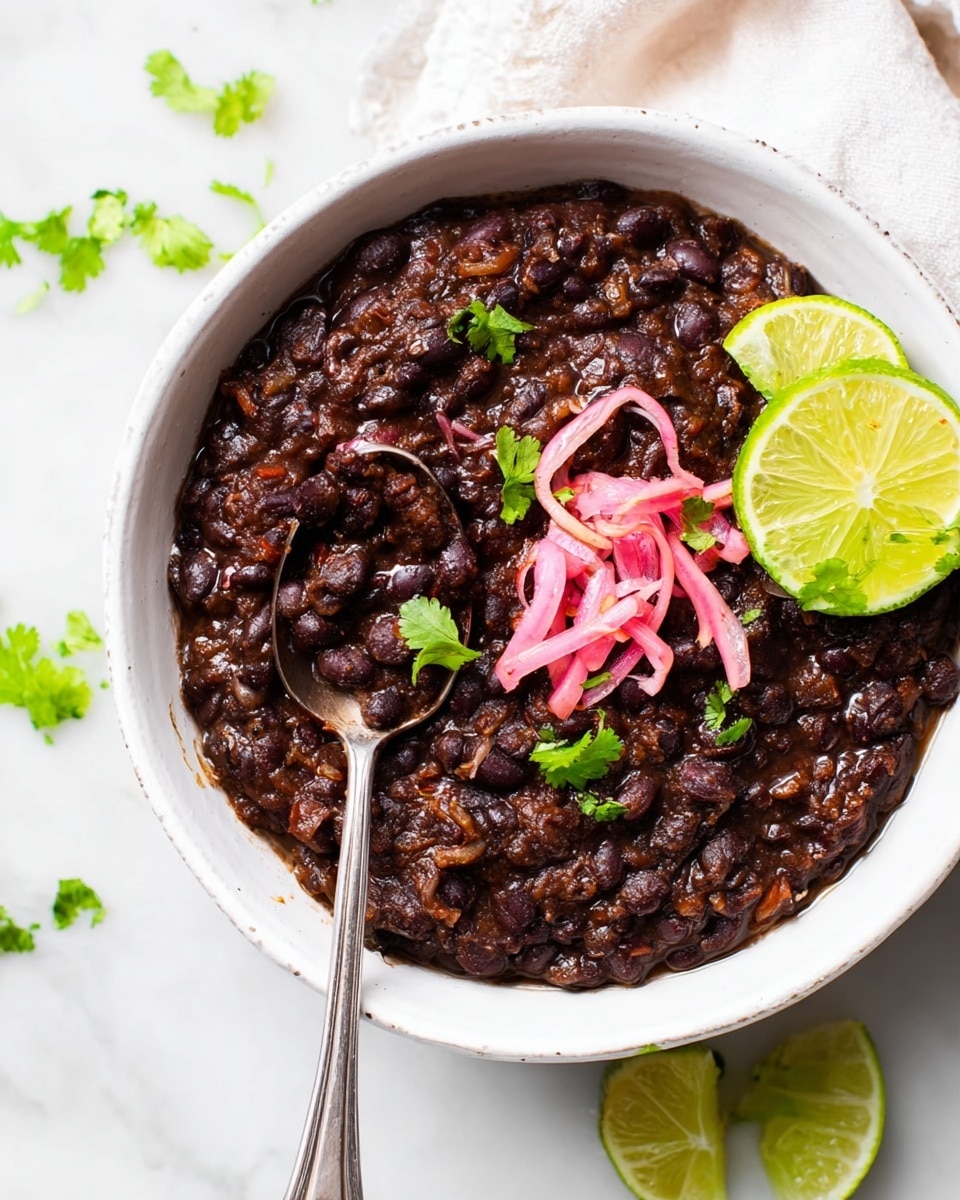 A white bowl filled with a thick, dark brown-black mix of cooked black beans, with a slightly chunky texture. On top, there are a few small sprigs of bright green cilantro leaves and thin, pale pink strips of pickled onions. Two lime wedges with bright green skin and pale yellow flesh rest on one side of the bowl. A silver spoon, scooping some of the beans, is placed inside the bowl. The bowl sits on a white marbled surface with scattered lime wedges and cilantro leaves visible around it. photo taken with an iphone --ar 4:5 --v 7