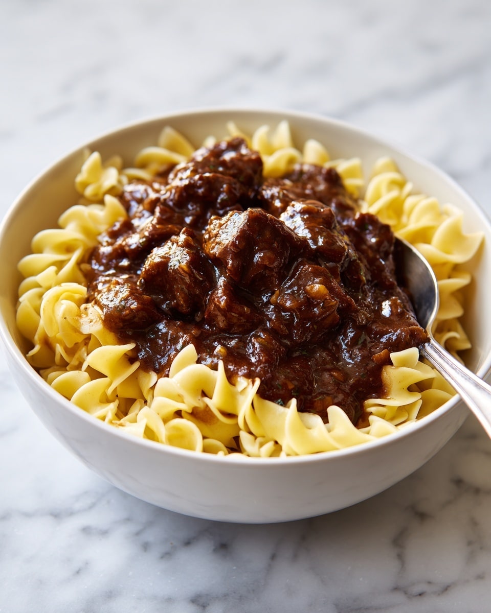 A white bowl filled with three layers: the bottom layer is light yellow egg noodles with ruffled edges, they look soft and slightly curled; the top layer has dark brown beef chunks covered in a thick, glossy brown gravy, the meat looks moist and tender; a silver spoon rests on the right edge of the bowl, slightly submerged in the noodles and gravy. The bowl is placed on a white marbled surface. photo taken with an iphone --ar 4:5 --v 7