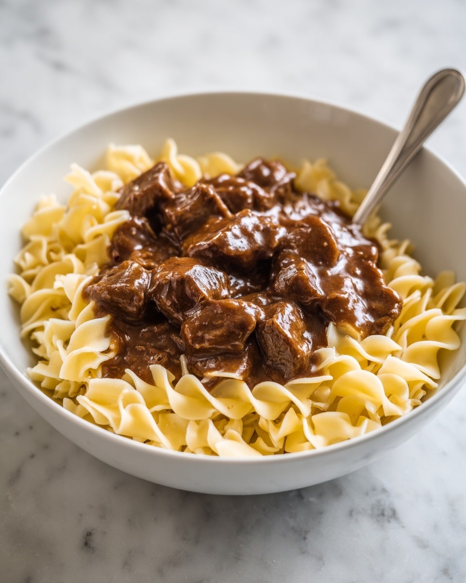 A white bowl holds a bed of yellow, wavy egg noodles forming the bottom layer with their soft, curly texture. On top, the second layer is composed of chunky beef stew with a thick, dark brown gravy that coats the tender pieces of meat. A silver fork rests at the edge of the bowl, partially submerged in the noodles and stew. The bowl sits on a white marbled surface. Photo taken with an iphone --ar 4:5 --v 7