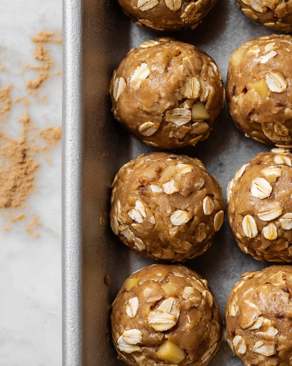 A close-up shot of several round cookie dough balls placed in a silver baking tray. Each ball shows a light brown dough base mixed with visible pieces of small, yellow apple chunks and scattered rolled oats on the surface, adding a slightly rough texture. The dough balls are closely set in a single layer, appearing soft and moist with a mix of smooth and textured areas caused by the oats and fruit pieces. The background features a white marbled surface with some specks of brown crumbs nearby. photo taken with an iphone --ar 4:5 --v 7