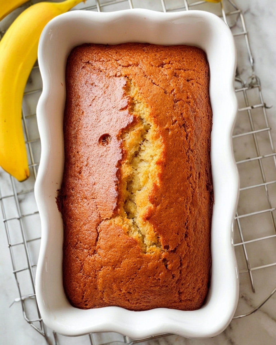 A rectangular loaf of golden brown banana bread is shown fresh out of a white ceramic baking dish with wavy edges. The top of the bread has a cracked, slightly uneven surface with a lighter yellow crumb visible through the cracks. The bread is thick and occupies the entire baking dish, showing a soft but firm texture. The dish rests on a white marbled surface with a baking rack partially visible beneath. Two ripe yellow bananas lie near the top corners of the image. Photo taken with an iphone --ar 4:5 --v 7