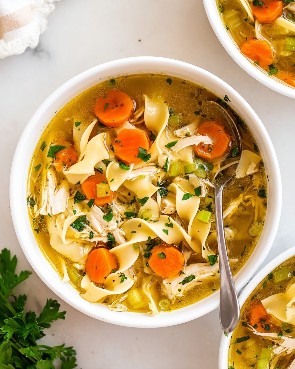 A white bowl filled with clear golden broth soup, containing large flat noodles that curl gently within the liquid. Bright orange carrot slices and shredded white chicken pieces are scattered evenly throughout. Small pieces of green celery and freshly chopped green herbs float on top, adding bursts of color. A silver spoon rests inside the bowl, and part of another similar bowl is seen to the side. The setting is a white marbled surface with a bit of green parsley nearby. Photo taken with an iphone --ar 4:5 --v 7