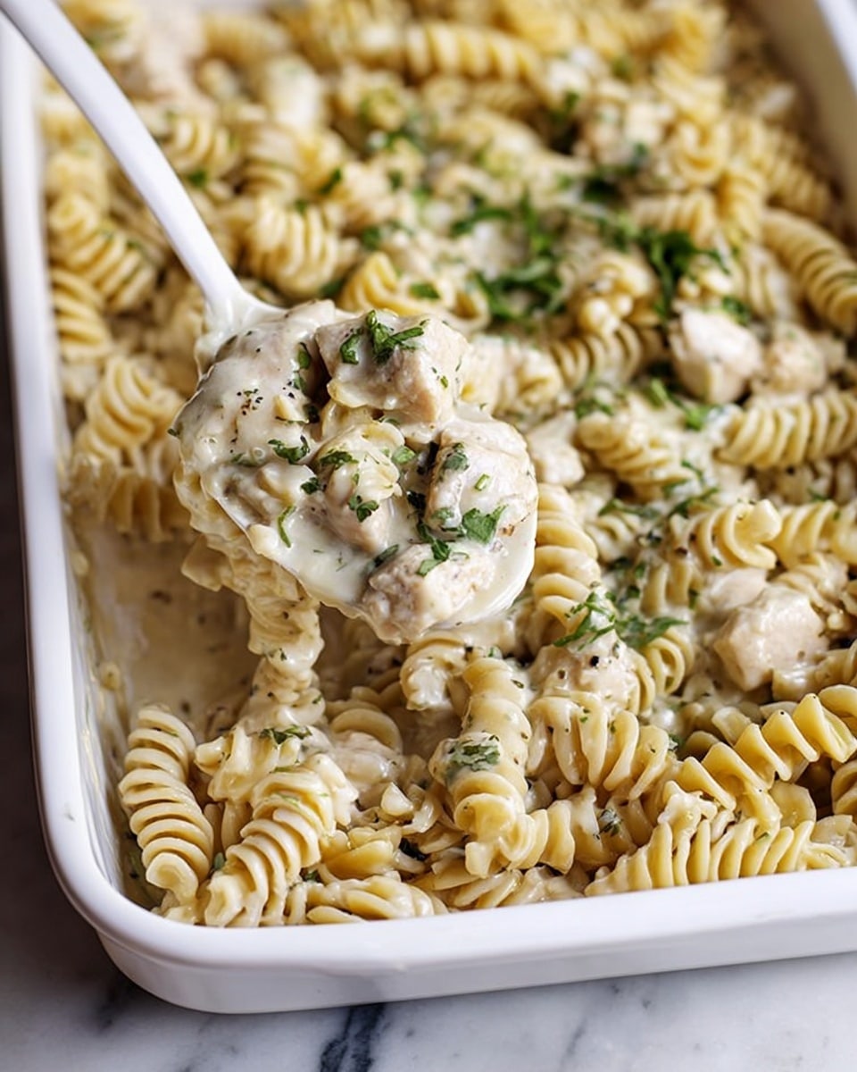 The image shows a white rectangular baking dish filled with rotini pasta mixed with chicken pieces. The pasta is light beige and twisted in shape, coated with a creamy sauce flecked with green herbs. The chicken chunks are white, tender-looking, and mixed evenly throughout the pasta. A white spoon is lifting a portion of the pasta and chicken from the dish, showing the close texture of the meal. The background is a white marbled surface, and there is a blurred bowl of green herbs in the background. photo taken with an iphone --ar 4:5 --v 7