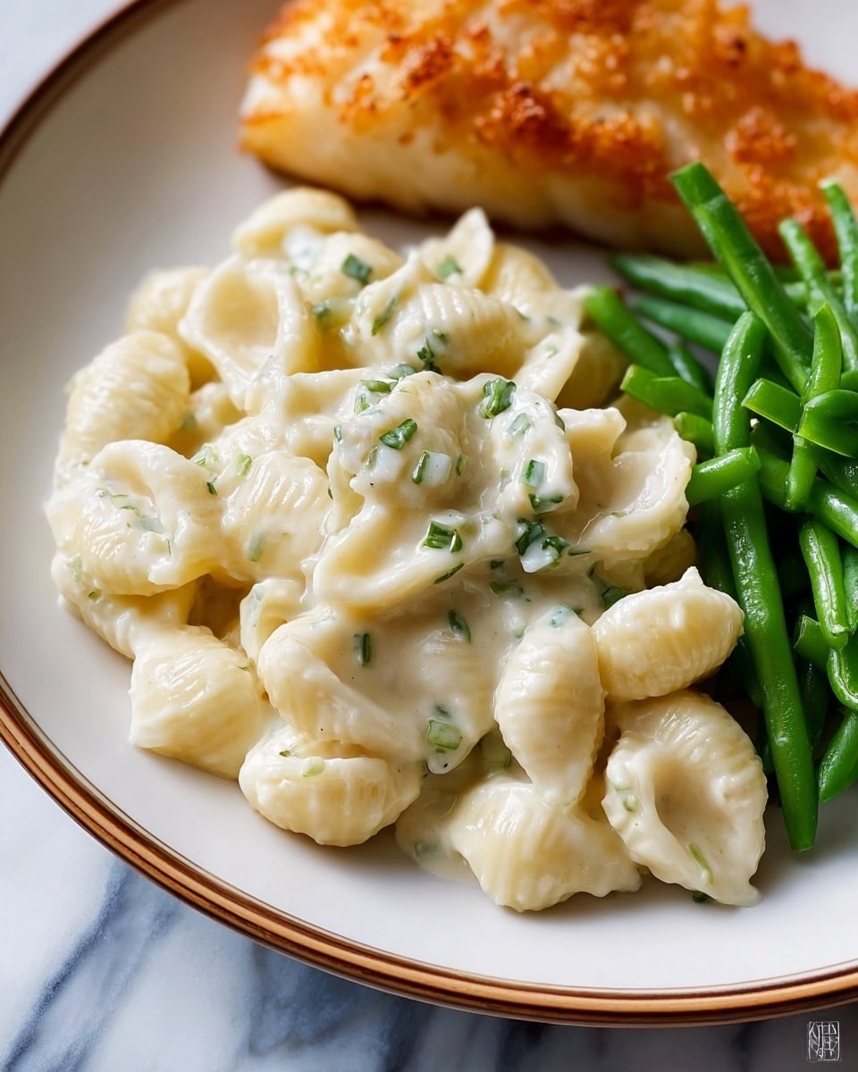 A close-up of a white plate with three main parts: in the center is a pile of creamy white pasta shells covered in a smooth, thick white sauce with small green herb bits; to the right are several bright green steamed green beans with a shiny, fresh texture; at the top of the plate is a piece of golden-brown cooked fish with a crispy texture. The plate sits on a white marbled surface. photo taken with an iphone --ar 4:5 --v 7