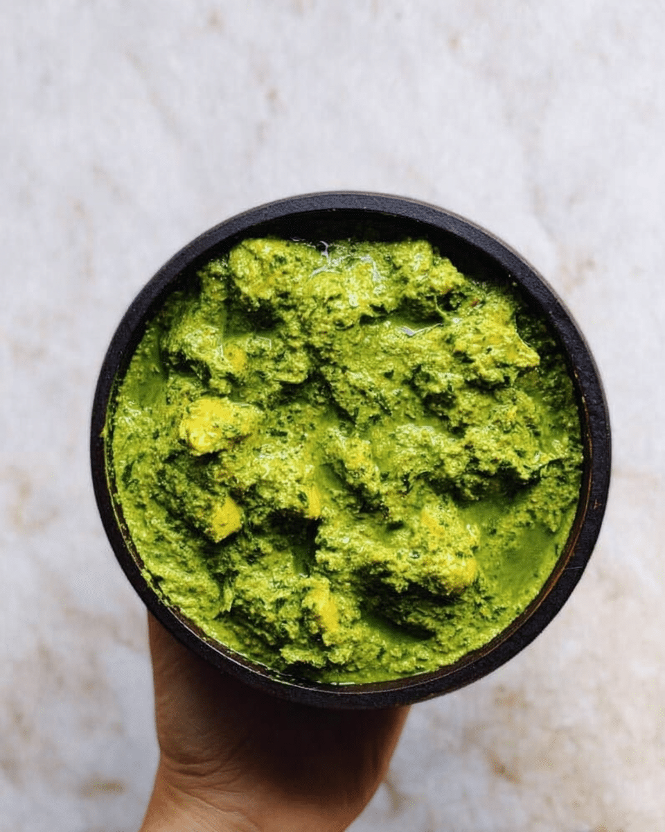 A top view of a round black bowl filled with a bright green thick textured dish with small yellowish-green chunks evenly spread throughout. The bowl is held by a woman's hand from the bottom center, above a white marbled surface. The green dish looks fresh and vibrant with a slightly rough texture showing the mix of ingredients. photo taken with an iphone --ar 4:5 --v 7