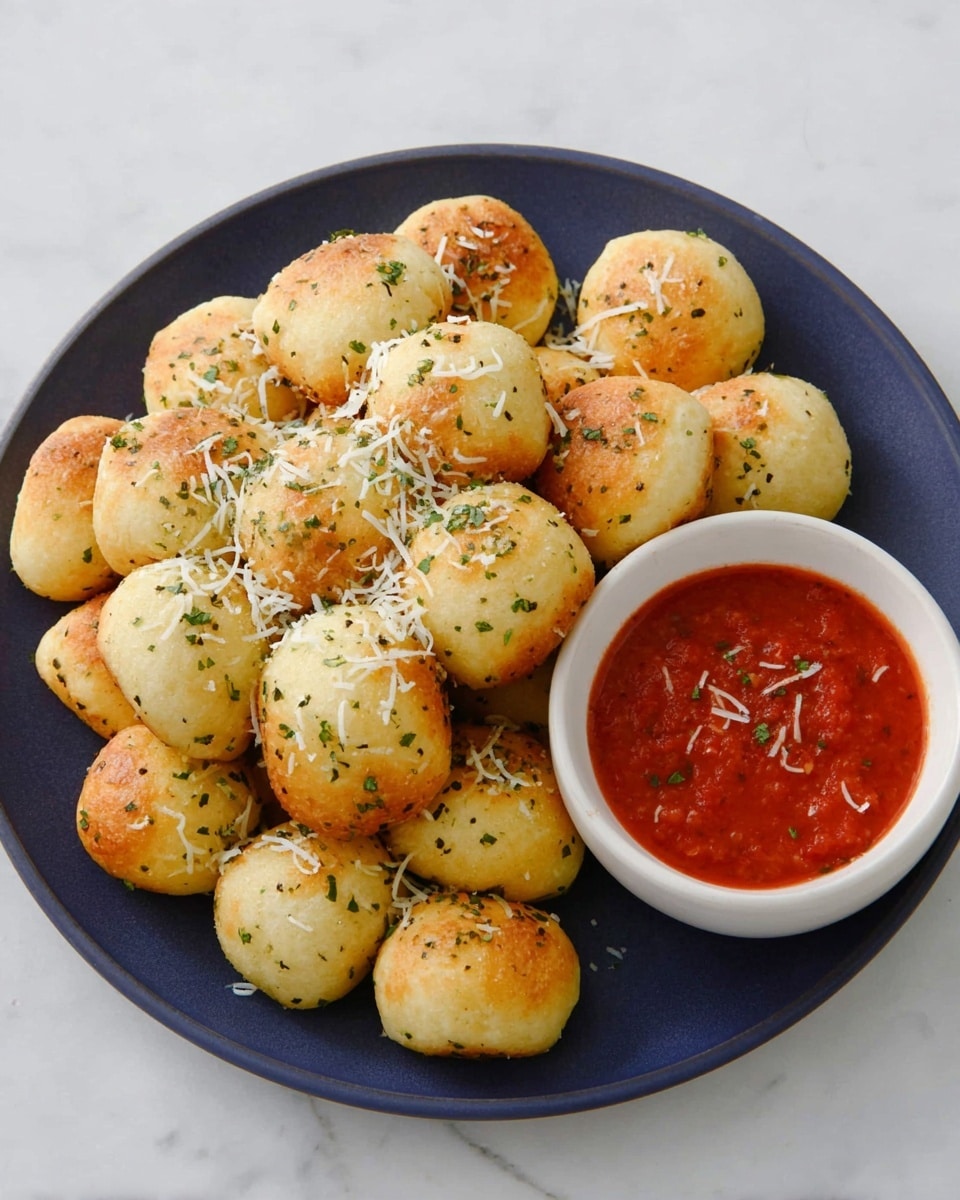 A dark blue round plate filled with many small golden-brown dough balls sprinkled with white grated cheese and green herbs, showing a soft, slightly shiny texture; to the right side of the plate, a small white bowl holds bright red marinara sauce with some bits of herbs and grated cheese on top; the plate is placed on a white marbled surface, creating a clean and fresh look. photo taken with an iphone --ar 4:5 --v 7