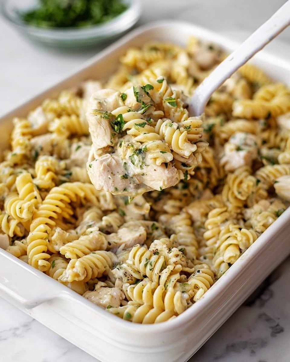 The image shows a white rectangular dish filled with creamy pasta made of spiral rotini noodles mixed with pieces of cooked chicken. The pasta is coated in a creamy white sauce with visible specks of green herbs and black pepper. Small bits of chopped green herbs are scattered throughout, adding color contrast. A white spoon is lifting a portion of the pasta, highlighting the smooth sauce and tender chicken chunks. The dish is placed on a white marbled surface. photo taken with an iphone --ar 4:5 --v 7