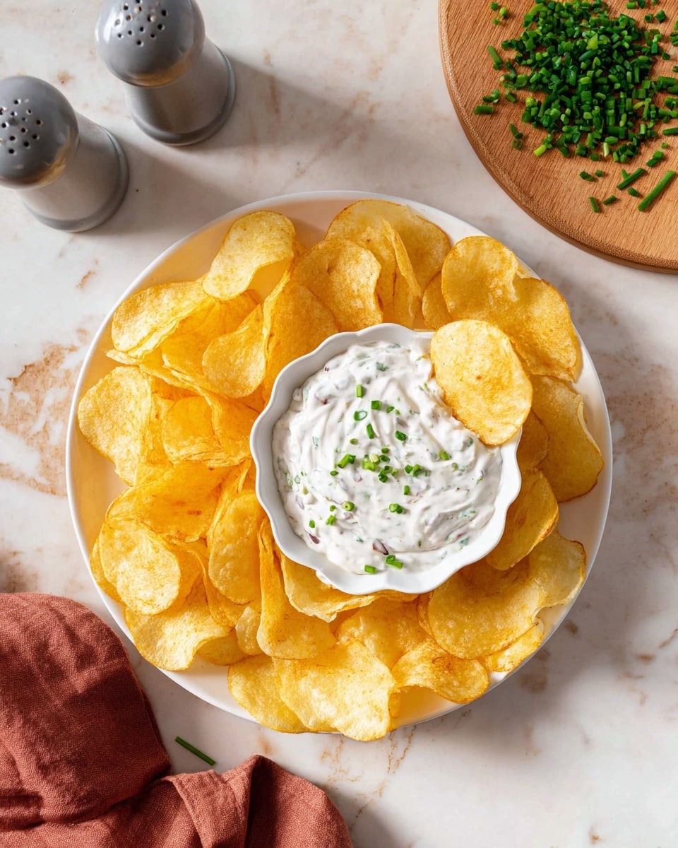 A white plate holds a pile of golden-yellow potato chips arranged in a loose circle around a small white scalloped bowl filled with creamy white dip. The dip is mixed with small bits of dark ingredients and is topped with small green chive pieces scattered evenly on the surface. One potato chip is partially dipped inside the bowl, resting on the edge. The plate is set on a white marbled surface with a round wooden board with chopped green chives in the top right corner, a pair of grey salt and pepper shakers to the top left, and a folded rust-colored cloth partially visible at the bottom left. Photo taken with an iphone --ar 4:5 --v 7