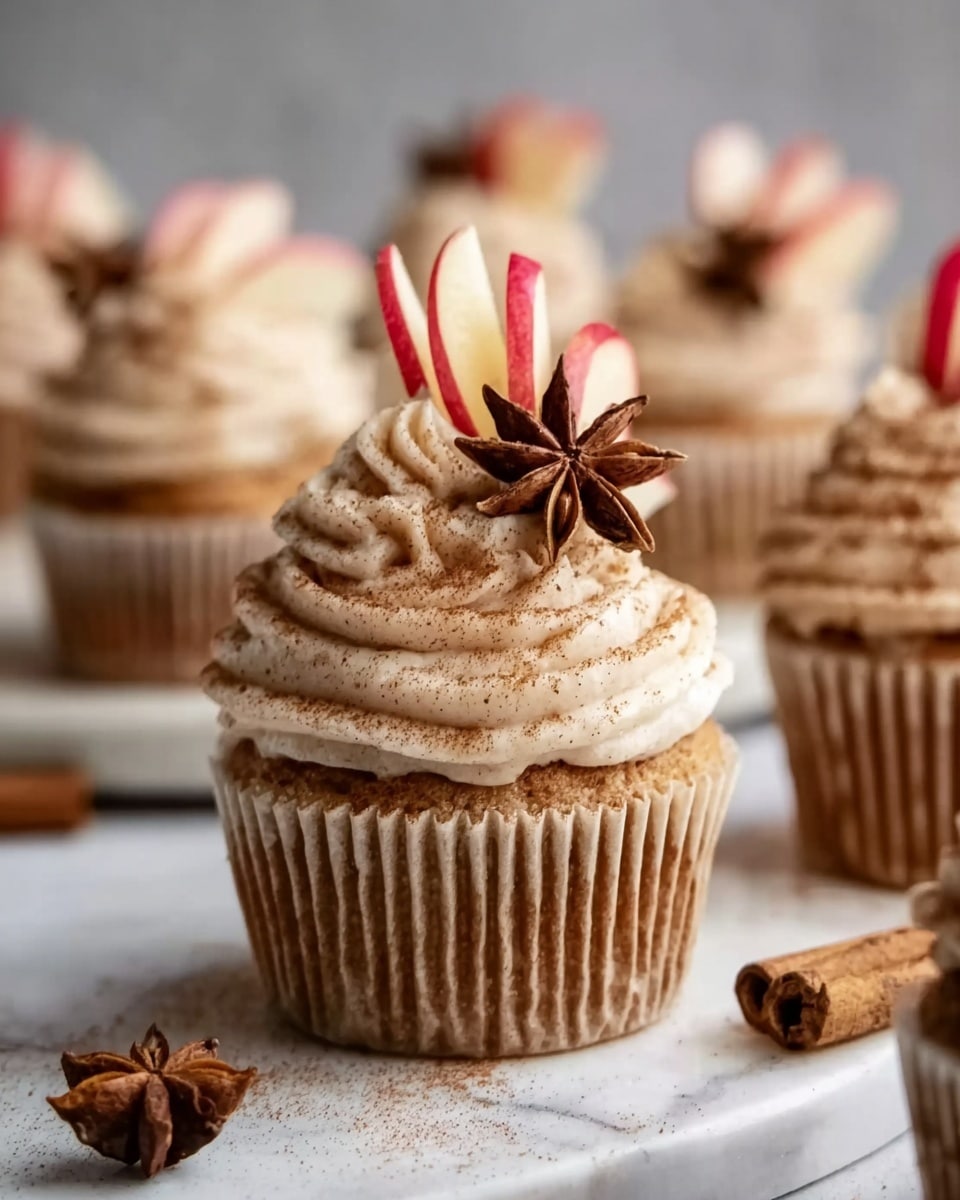 The image shows close-up cupcakes on a white marbled surface. Each cupcake has a light brown base with ridged texture from the cupcake liner, topped with a thick swirl of light beige frosting sprinkled with cinnamon powder. On top of the frosting, there is a thin slice of red apple and a few star anise pieces, adding decoration. The background is softly blurred with more cupcakes visible, creating a warm and cozy feeling. Photo taken with an iphone --ar 4:5 --v 7