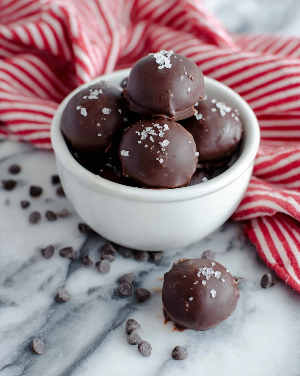 A small white bowl holds about seven round chocolate balls with a smooth, dark brown chocolate coating, sprinkled lightly with coarse sea salt flakes on top. Two chocolate balls lie outside the bowl on a white marbled surface, surrounded by scattered dark chocolate chips. A red and white striped cloth is casually placed behind and to the side of the bowl, adding a cozy touch to the scene. The chocolate balls appear rich and glossy, showing a few small dimples and bumps from the coating. Photo taken with an iphone --ar 4:5 --v 7