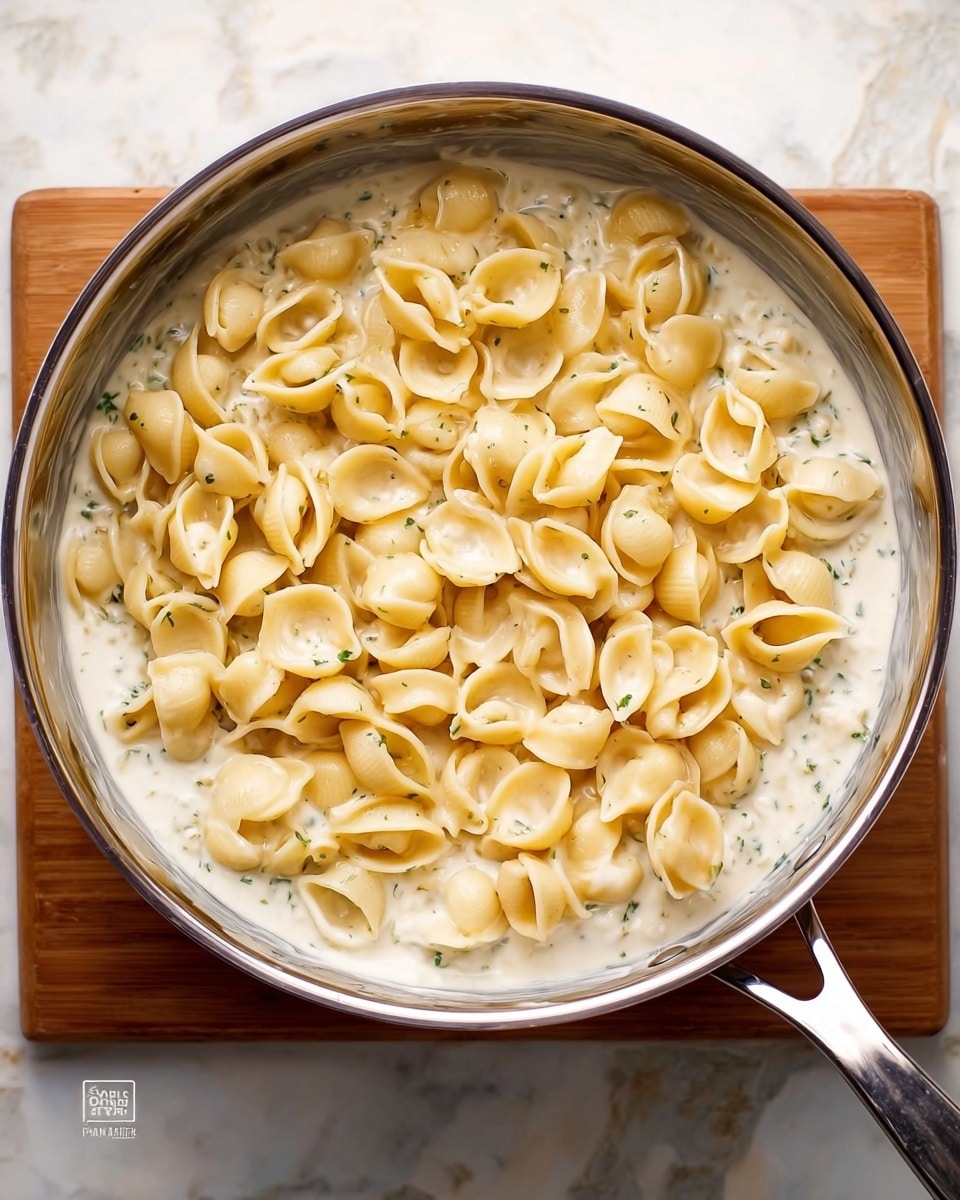 A shiny metal pan placed on a white marbled surface holds a dish with two clear layers visible; the bottom layer is a creamy white sauce with some small green herb bits, and the top layer is light beige conchiglie pasta shells evenly spread over the sauce, showing their curved and hollow shapes. The pan has a handle on the right side. photo taken with an iphone --ar 4:5 --v 7