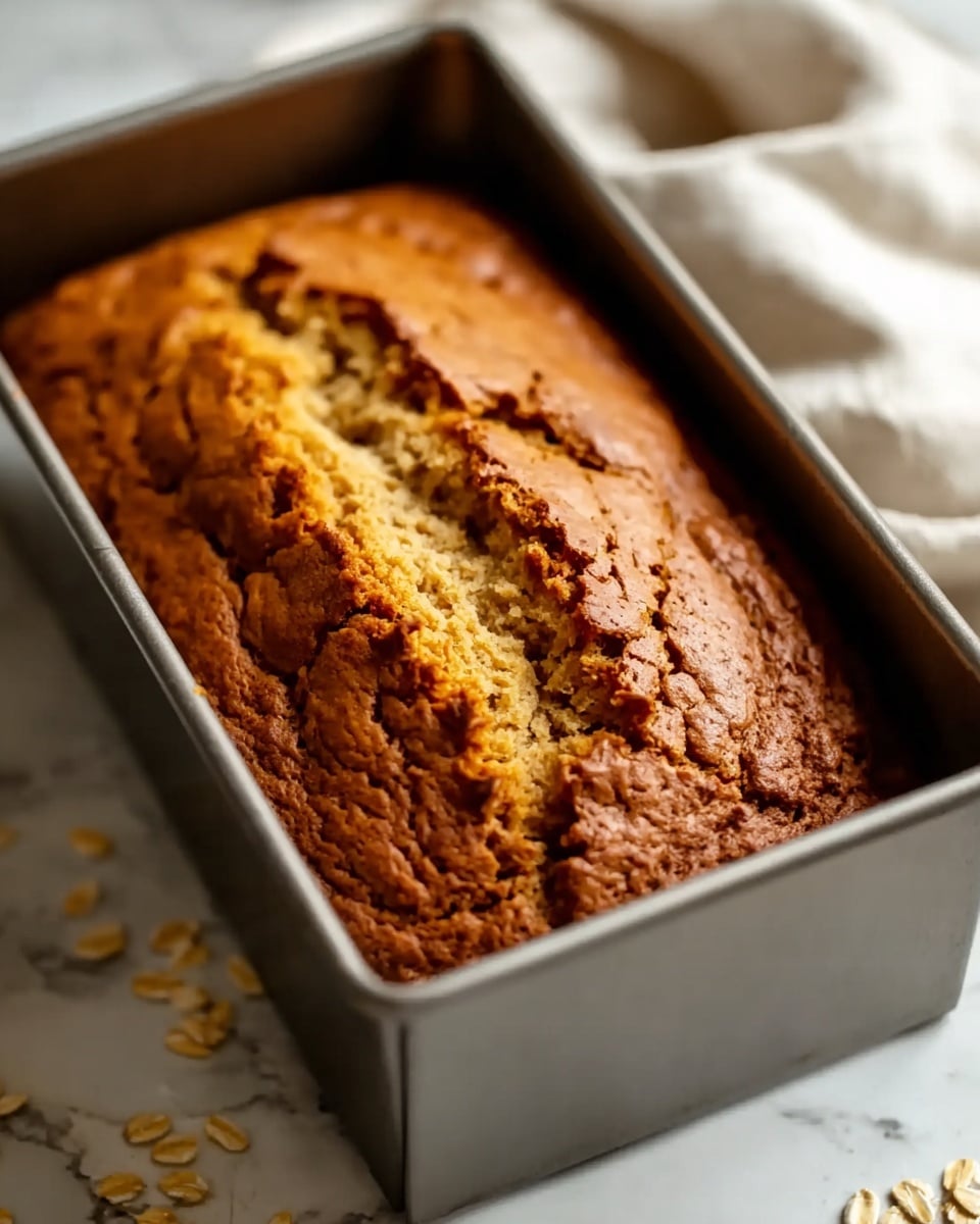 A golden brown loaf cake with a slightly cracked top showing a lighter, moist inside layer in the middle, resting inside a silver baking pan on a white marbled surface; the cake surface has a rough, textured appearance with darker brown edges and a small piece flaked off near the top right corner; the background includes a blurred white cloth and some scattered oats on the side. photo taken with an iphone --ar 4:5 --v 7