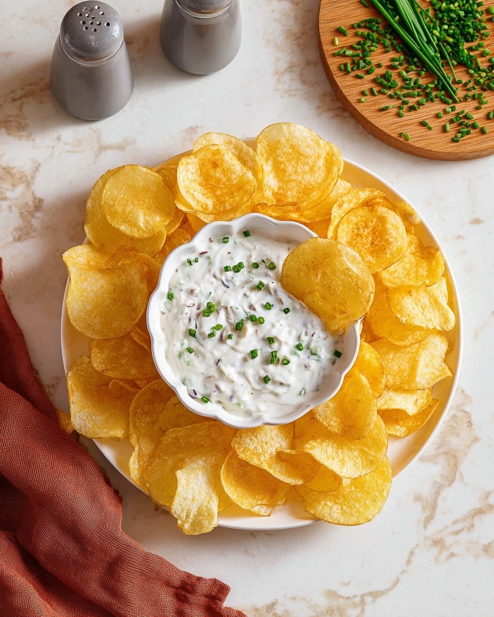 The image shows a white scalloped bowl filled with creamy white dip with visible small pieces of mushrooms mixed in, topped with bright green chopped chives. The bowl is centered on a white plate surrounded by yellow potato chips. In the background, there is a wooden board with more chopped chives and a black and silver pepper grinder on a white marbled surface. photo taken with an iphone --ar 4:5 --v 7