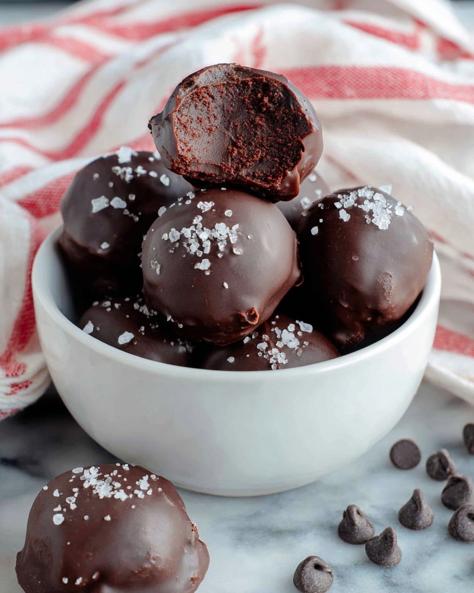 The image shows a white bowl filled with six round dark chocolate truffles, each coated smoothly with a shiny chocolate layer and sprinkled lightly with white sea salt on top. One truffle at the top of the bowl is bitten, revealing a dense, rich, dark chocolate fudge center inside. Around the bowl on a white marbled surface, there are three more truffles, each with a shiny chocolate coating and a small sprinkle of sea salt. Small round dark chocolate chips are scattered loosely near the truffles. A white cloth with red stripes partially covers the background adding a soft contrast. photo taken with an iphone --ar 4:5 --v 7