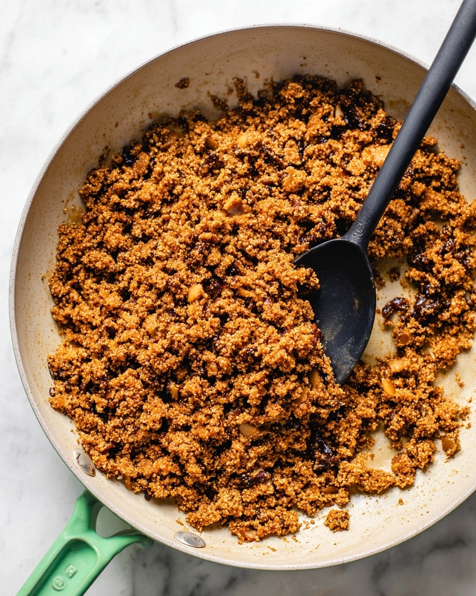 The image shows a close-up of cooked ground mixture in a light-colored pan with a green handle. The mixture is crumbly and uneven in texture, with small bits of darker and lighter brown pieces mixed throughout, likely cooked mushrooms or similar ingredients. A black spoon is resting in the pan, partially stirring the mixture. The pan is placed on a white marbled surface. photo taken with an iphone --ar 4:5 --v 7
