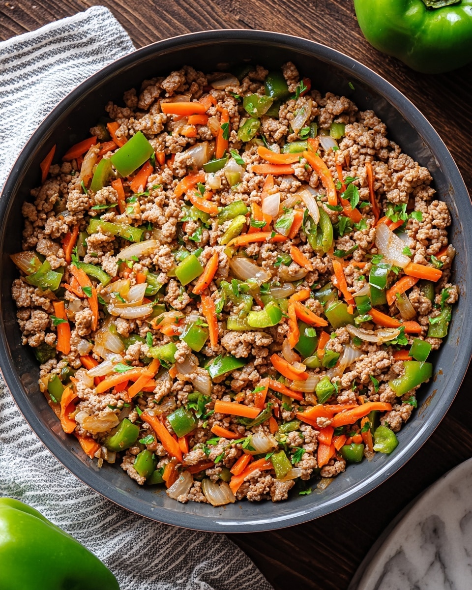 A dark gray round pan filled with a cooked dish of ground meat mixed with colorful vegetables. The dish has three main layers: a base of browned, crumbly ground meat; scattered chopped green bell peppers and orange carrot sticks adding bright color and crunch; and pieces of translucent cooked onions mixed evenly throughout. Small bits of fresh green herbs are sprinkled on top, giving a fresh touch. The pan sits on a dark wooden surface with a white and gray striped cloth visible at the bottom left edge and a green bell pepper positioned on the right side on a white marbled texture. photo taken with an iphone --ar 4:5 --v 7