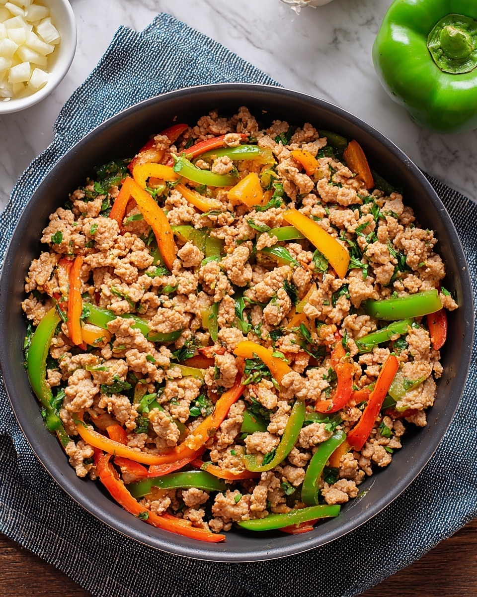 A dark round pan filled with a cooked mixture of ground meat and sliced bell peppers in three colors: green, red, and yellow, all mixed with small bits of green herbs scattered evenly. The ground meat is light brown, crumbly, and sits at the base and middle of the pan, mingled with the soft, slightly shiny strips of bell peppers. This pan rests on a blue-gray striped cloth over a white marbled surface. A small white dish with chopped garlic pieces is nearby, and a bright green bell pepper is visible in the background. photo taken with an iphone --ar 4:5 --v 7