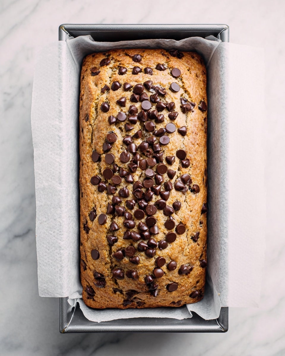 A single rectangular loaf of golden-brown bread is shown inside a metal baking pan lined with white parchment paper, with the edges of the paper extending slightly beyond the pan. The top of the bread is textured with a light crust, and there is a generous spread of dark brown chocolate chips scattered thickly across the center area of the loaf. The baking pan is placed on a white marbled surface that provides a clean and soft background. Photo taken with an iphone --ar 4:5 --v 7