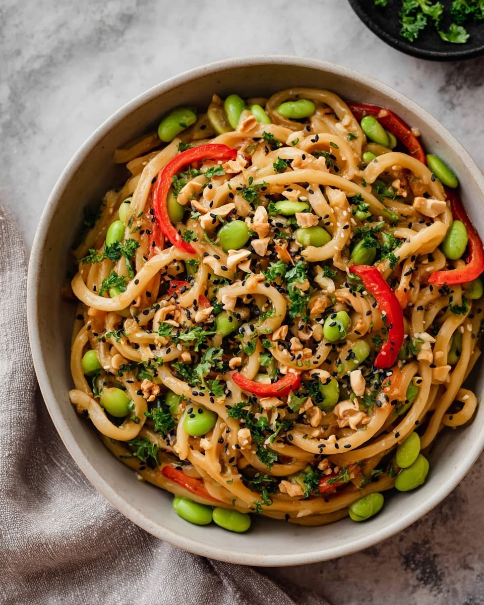 A deep white bowl holds a colorful noodle dish with three main layers: the bottom layer is thick, light brown udon noodles that are twisted and shiny; the middle layer has bright green edamame beans and sliced red bell peppers spread evenly; the top layer is sprinkled with fresh green herbs, chopped green onions, small white and black sesame seeds, and crushed nuts, adding texture and color. The bowl sits on a white marbled texture surface with some sesame seeds scattered around. In the background, there is a partial view of a white bowl with leafy green vegetables. photo taken with an iphone --ar 4:5 --v 7