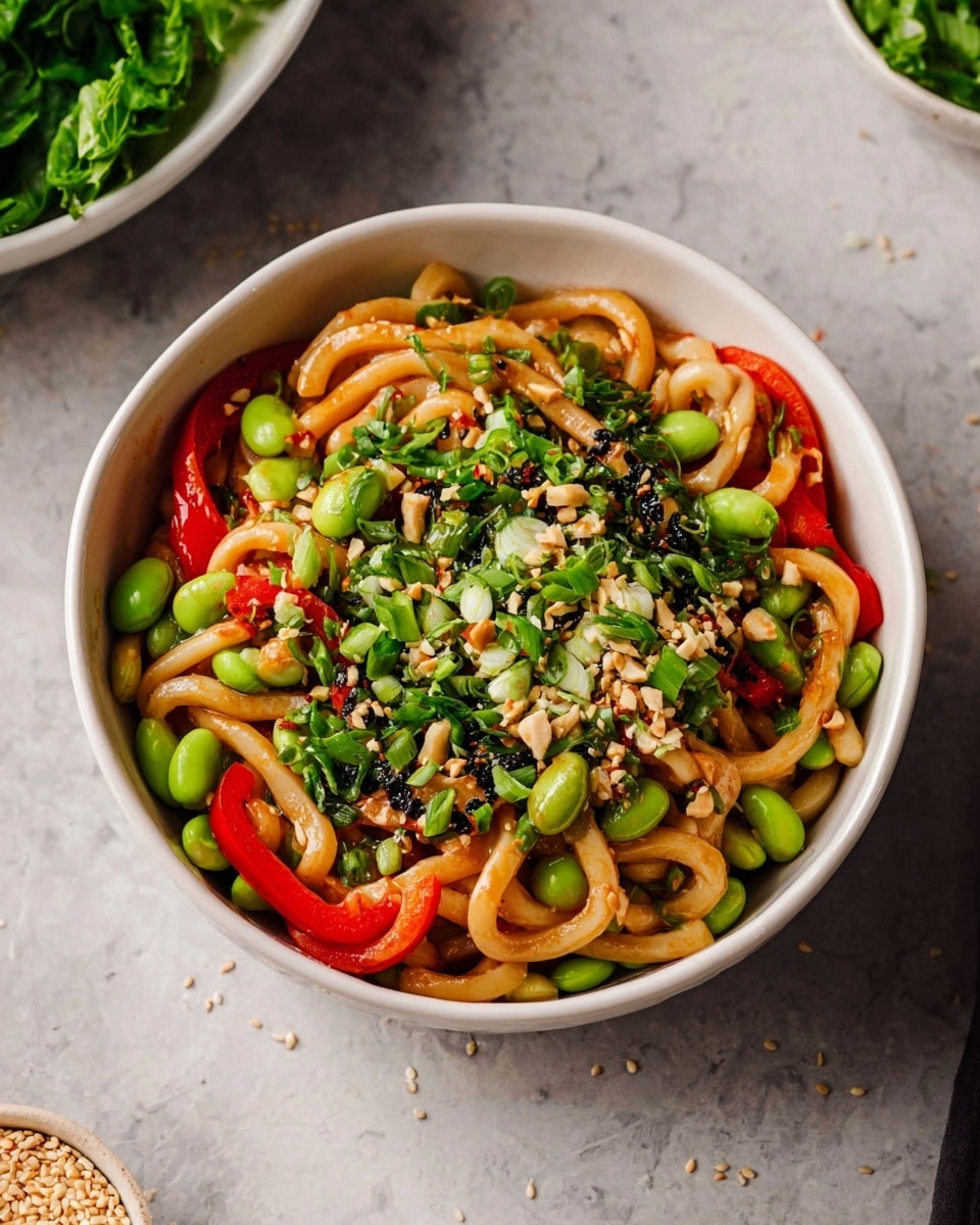 A white bowl holds thick light tan noodles twisted and curled, mixed with bright green edamame beans scattered throughout. Thin, curved strips of red bell pepper add vibrant color on top and around the noodles, while finely chopped dark green herbs are evenly spread across the dish. Small black sesame seeds are sprinkled over, adding texture contrast, and bits of chopped nuts are visible, giving a varied look. The bowl is set on a surface with a white marbled texture, next to a soft light gray cloth. Photo taken with an iphone --ar 4:5 --v 7