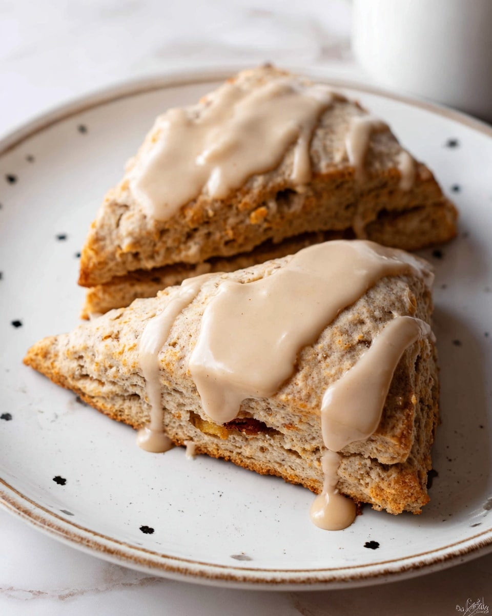 Two thick, triangular scones with a light brown color and visible crumbly texture are placed on a white plate with small black dots near the edge. Each scone is topped with a smooth, creamy beige glaze that drips slightly down the sides. The scones have layers that show a soft inside with some pieces of fruit or nuts visible at the bottom layer. The plate sits on a white marbled surface. photo taken with an iphone --ar 4:5 --v 7