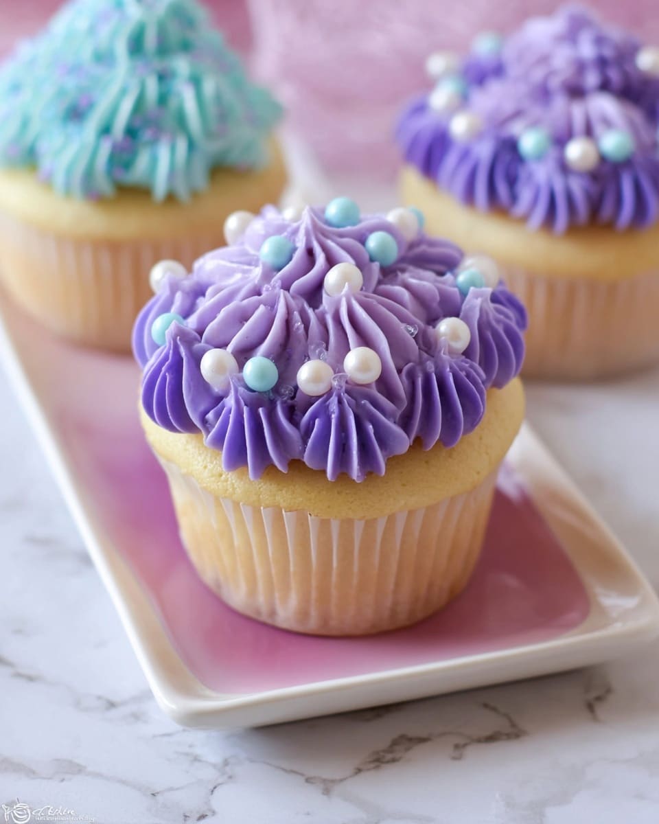 A vanilla cupcake with a pale yellow base is topped with eight purple frosting dollops, arranged in a circle with a slightly pointed texture. Scattered among the frosting are small, round white and light blue sugar pearls, adding a shiny contrast. The cupcake is in a translucent purple paper liner. It sits on a white rectangular plate with a pale pink center, placed over a white marbled surface. In the background, two similar cupcakes with purple and blue frosting can be seen slightly out of focus. Photo taken with an iphone --ar 4:5 --v 7