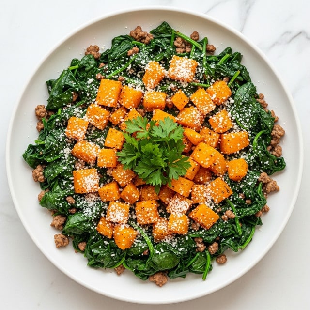 The image shows a white plate filled with a dish made of several layers: at the bottom are cooked spinach leaves that are dark green and soft, mixed throughout with small browned ground meat bits. On top of this, there are bright orange cubes of roasted squash scattered evenly. A light sprinkle of grated white cheese covers the whole plate, adding fine texture and contrast. The dish is garnished in the center with a small bunch of fresh green parsley. The plate sits on a white marbled surface. Photo taken with an iphone --ar 4:5 --v 7