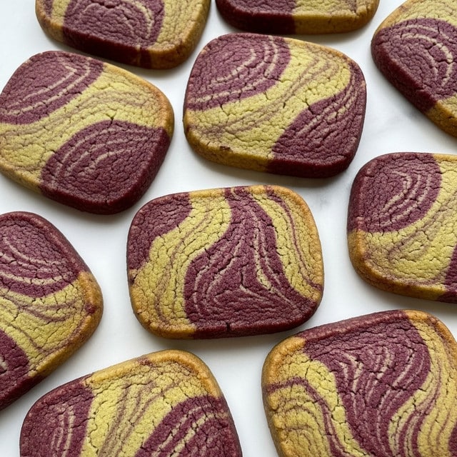 The image shows many square-shaped cookies scattered on a white marbled surface. Each cookie has a smooth texture with a colorful pattern of two main colors: purple and green. The patterns include lines, swirls, and shapes that alternate between the two colors, creating a marble-like effect. The cookies have a slightly golden-brown edge, indicating they are baked. There are no additional items or decorations in the image. photo taken with an iphone --ar 4:5 --v 7