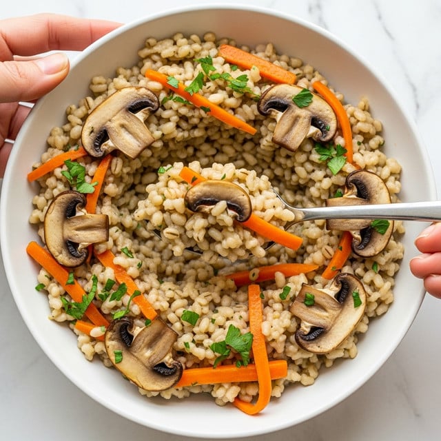 A close-up of creamy mushroom barley soup in a large white bowl on a white marbled surface, showing two main layers: the base is a thick, light beige broth mixed with soft barley grains, small diced orange carrots, and finely chopped green herbs sprinkled on top. Scattered throughout the soup are sautéed dark brown mushroom slices with visible texture and some glistening oil drops. A silver spoon, held by a woman's hand, scoops up a portion revealing the chunky mix of barley, mushrooms, carrots, and herbs all combined in the creamy broth. photo taken with an iphone --ar 4:5 --v 7