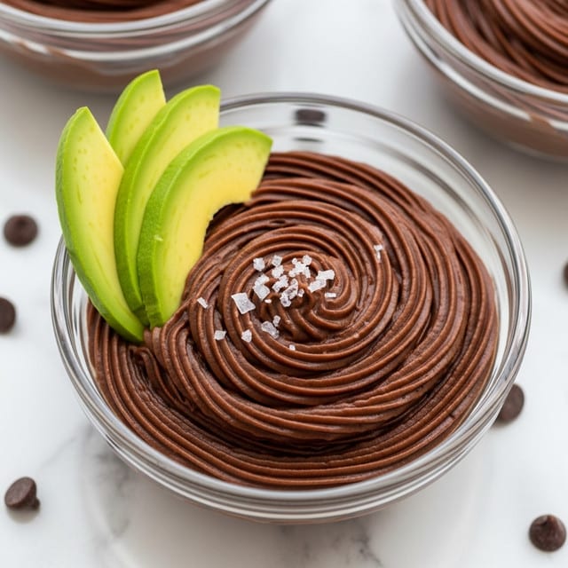 The image shows a clear glass cup filled with two layers of smooth, dark brown chocolate mousse. On top, there is a swirl pattern that adds texture to the mousse. Three thin slices of bright green avocado are placed on one side of the mousse surface, slightly overlapping each other. A small pinch of coarse white salt is sprinkled over the mousse, right next to the avocado. The cup is placed on a white marbled surface, scattered with a few dark brown chocolate chips around it. In the background, two similar glasses filled with chocolate mousse are partially visible. photo taken with an iphone --ar 4:5 --v 7