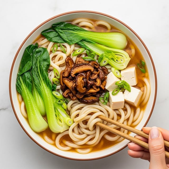A white bowl filled with a noodle soup showing three main layers: the bottom broth layer which is light brown and clear, floating thick white noodles creating a tangled base, and on top, a pile of dark brown cooked mushrooms. Around the edges, pieces of bright green bok choy and white tofu cubes with smooth surfaces float along with sliced green onions. A woman's hand holds light wood chopsticks picking up some noodles near the bottom right. The bowl is on a white marbled surface with a small white dish of orange sauce nearby. Photo taken with an iphone --ar 4:5 --v 7
