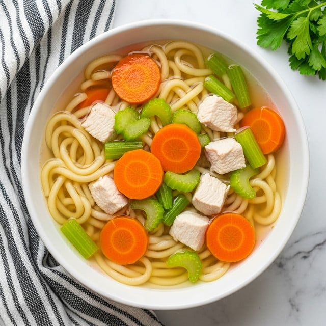 A white bowl filled with chicken noodle soup is placed on a white marbled surface with a striped cloth nearby. The soup has a clear broth with yellowish noodles that are long and thick, forming the base layer. On top are evenly spread round carrot slices in bright orange and small chunks of white cooked chicken. Also mixed among the layers are pieces of light green celery adding a fresh crunch. Small green parsley leaves float lightly on the surface, adding a touch of color contrast. The photo is taken from above, showing all details clearly. photo taken with an iphone --ar 4:5 --v 7