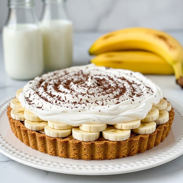 The image shows a tart with three visible layers, placed on a white plate with a textured edge. The bottom layer is a golden-brown crust with a slightly rough texture and visible fluted edges. Above this is a layer of evenly sliced bananas arranged in a circular pattern, their light yellow color contrasting with the crust. The top layer is a thick swirl of white whipped cream with delicate peaks, sprinkled with fine chocolate shavings that add a speckled dark brown color on top. In the background, there are two glass bottles filled with milk and a bunch of bananas, all set on a white marbled surface. Photo taken with an iphone --ar 4:5 --v 7