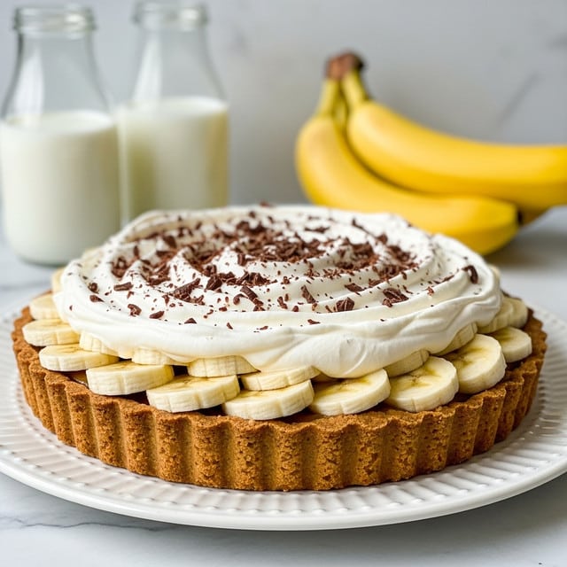 A tart with three visible layers is placed in the middle of a white plate on a white marbled surface. The bottom layer is a golden-brown crust with a slightly rough texture and fluted edges. On top of the crust, there is a layer of neatly arranged, evenly sliced banana pieces that are pale yellow with light brown specks. The top layer consists of a large, fluffy mound of white whipped cream, swirled with soft peaks and sprinkled with fine dark chocolate shavings. In the background, there are two glass bottles filled with a creamy white liquid and a bunch of bright yellow bananas. Photo taken with an iphone --ar 4:5 --v 7