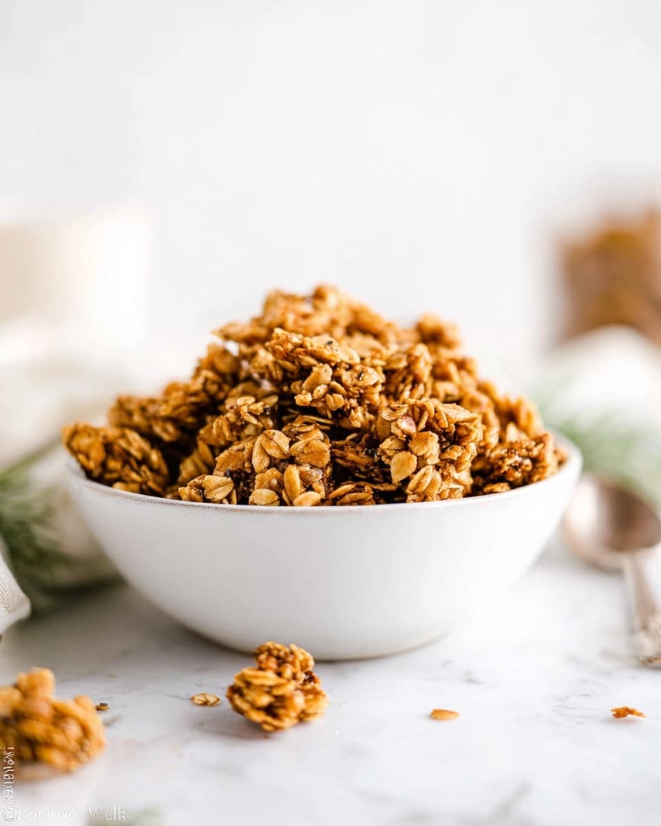 A white bowl filled with a large pile of crunchy granola clusters sits on a white marbled surface, with some loose granola pieces scattered around it. The granola is golden brown with visible oats and nuts, giving it a textured and crunchy look. The background is blurred with a soft white marbled texture, making the granola bowl the main focus. There is a silver spoon partially visible on the right side near the bowl. photo taken with an iphone --ar 4:5 --v 7