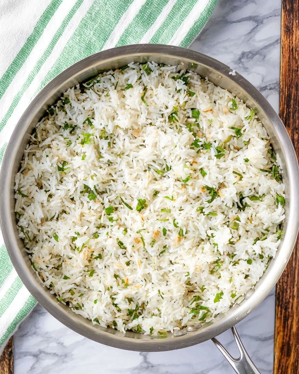 A shiny silver pan filled with one layer of cooked white rice mixed with small bits of green herbs and tiny light brown toasted pieces, giving the rice a slightly textured look. The pan rests on a white marbled surface, with a green and white striped cloth partially visible to the upper left. The rice appears fluffy and evenly spread inside the pan, with some herbs scattered mainly throughout. Photo taken with an iphone --ar 4:5 --v 7