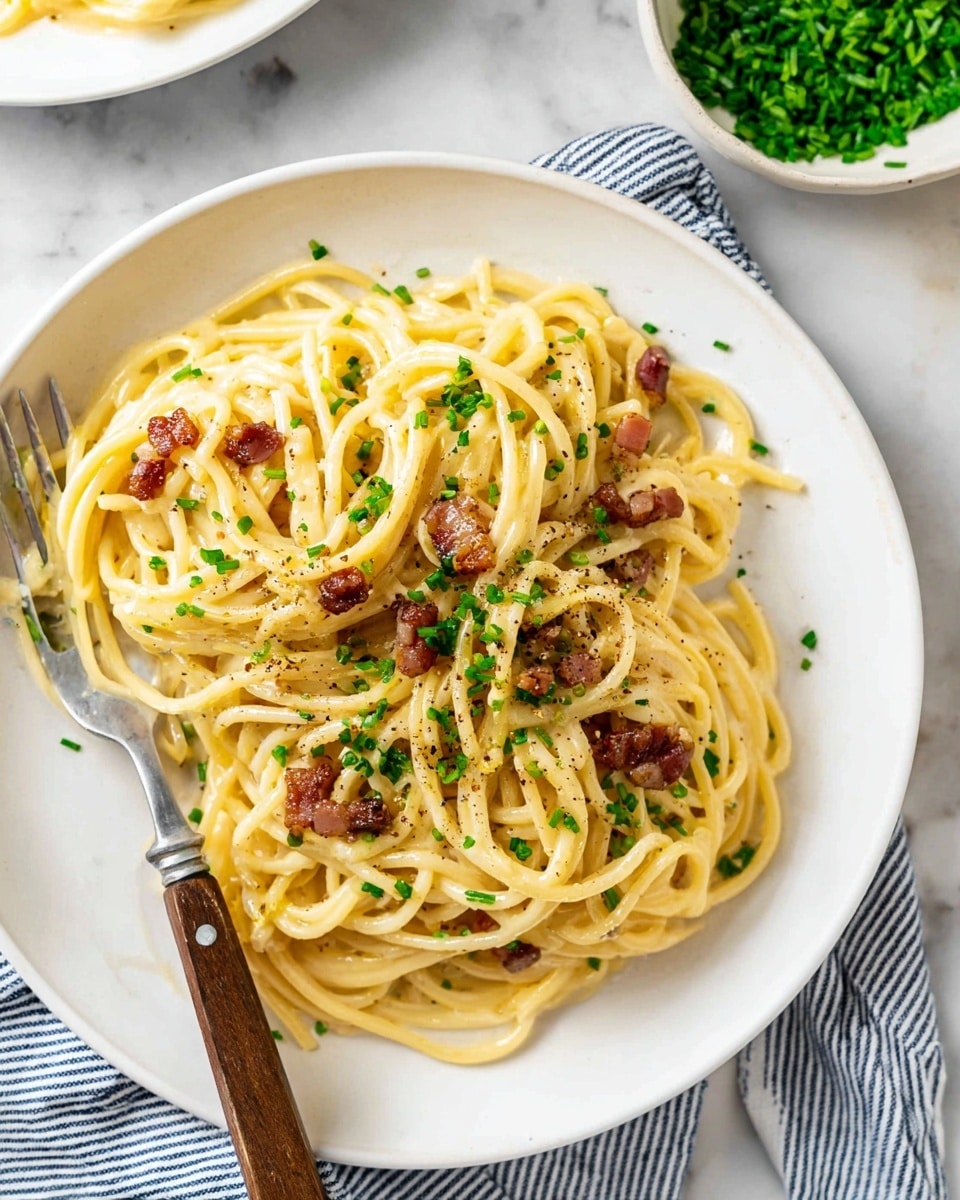 A white bowl holds spaghetti pasta with a creamy yellow sauce, mixed with small pieces of browned bacon scattered on top and throughout the noodles. Bright green chopped chives are sprinkled generously across the pasta, adding a fresh contrast. The pasta looks smooth and slightly glossy, with specks of black pepper visible on the surface. A fork with a wooden handle rests on the side of the bowl, partly inserted into the pasta. The bowl sits on a white marbled surface, with a small white bowl of extra chopped chives placed nearby. A blue and white striped cloth is also partially visible under the bowl. photo taken with an iphone --ar 4:5 --v 7