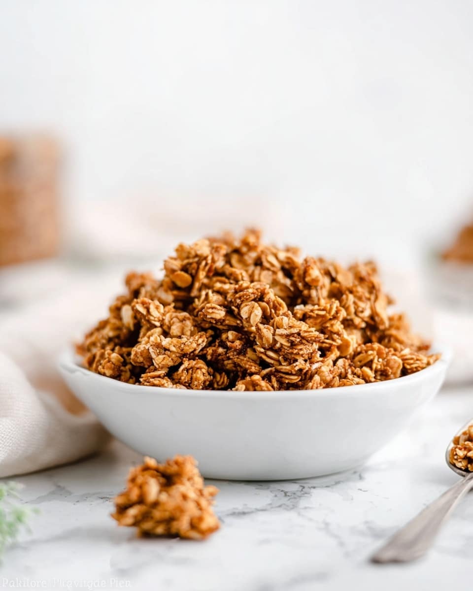 A close-up of a white shallow dish filled with a large pile of golden brown granola clusters. The granola pieces have a rough texture with visible oats and a slightly crispy look. One small cluster is placed outside the bowl on the white marbled surface, and a spoon is partially visible on the right side. The background is bright and softly blurred, emphasizing the granola in the foreground. photo taken with an iphone --ar 4:5 --v 7