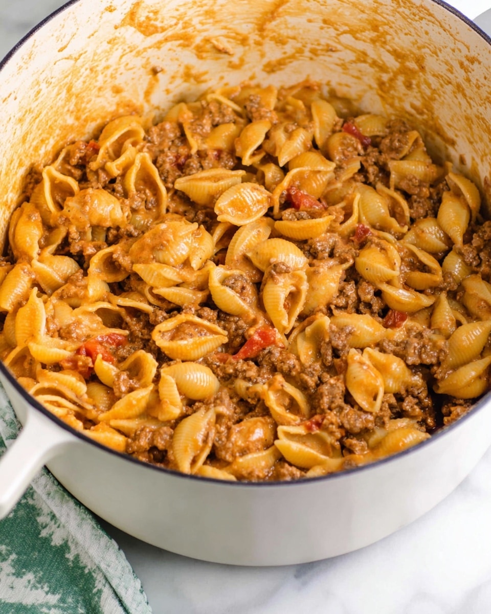 A large white pot filled with soft shell pasta coated in a creamy orange sauce, mixed with small pieces of browned ground meat and bits of red tomato, all evenly combined with a smooth texture; the inside walls of the pot show streaks of the sauce. The dish is thick and creamy with a rich, hearty appearance, sitting on a white marbled surface with a green-striped cloth partially visible. photo taken with an iphone --ar 4:5 --v 7