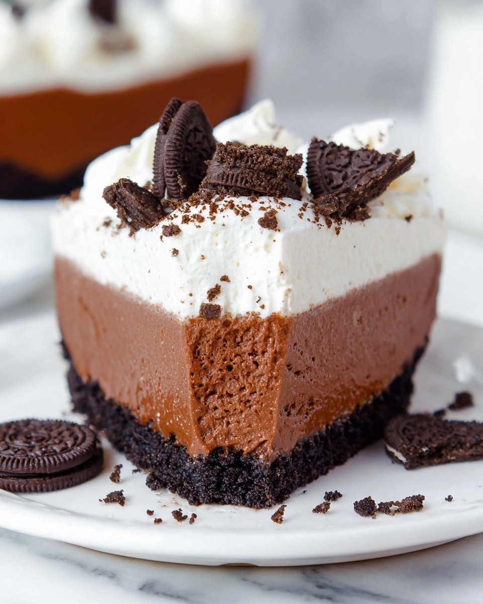 A slice of pie with three clear layers lies on a white plate over a white marbled surface. The bottom crust is dark, crumbly, and thick with a rough texture. The middle layer is smooth and dense chocolate brown, taking up the most space. The top layer is fluffy white cream, decorated with small dollops and topped with broken pieces of dark chocolate cookies scattered on and around the slice. The background shows other similar slices softly focused. Photo taken with an iphone --ar 4:5 --v 7