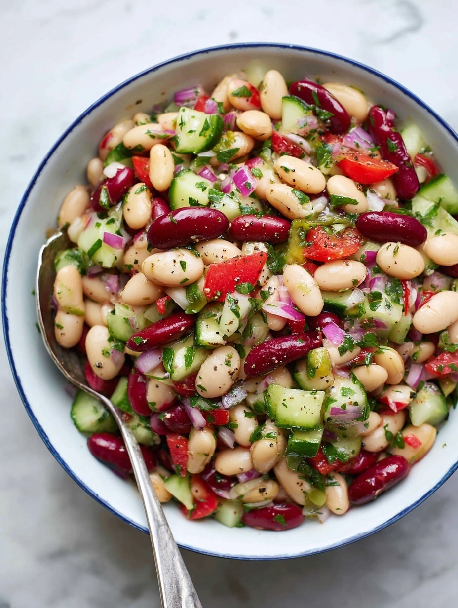 A close-up view of a colorful bean salad in a white bowl with a blue rim, showing three main layers mixed together: light beige chickpeas, red kidney beans, and white beans, combined with chopped green cucumber pieces and small red tomato chunks. There are finely chopped green herbs and bits of purple onion spread evenly throughout, with a sprinkling of black pepper on top. A silver serving spoon and fork rest inside the bowl on the left side, slightly covered by the salad. The whole scene is set on a white marbled surface. photo taken with an iphone --ar 4:5 --v 7