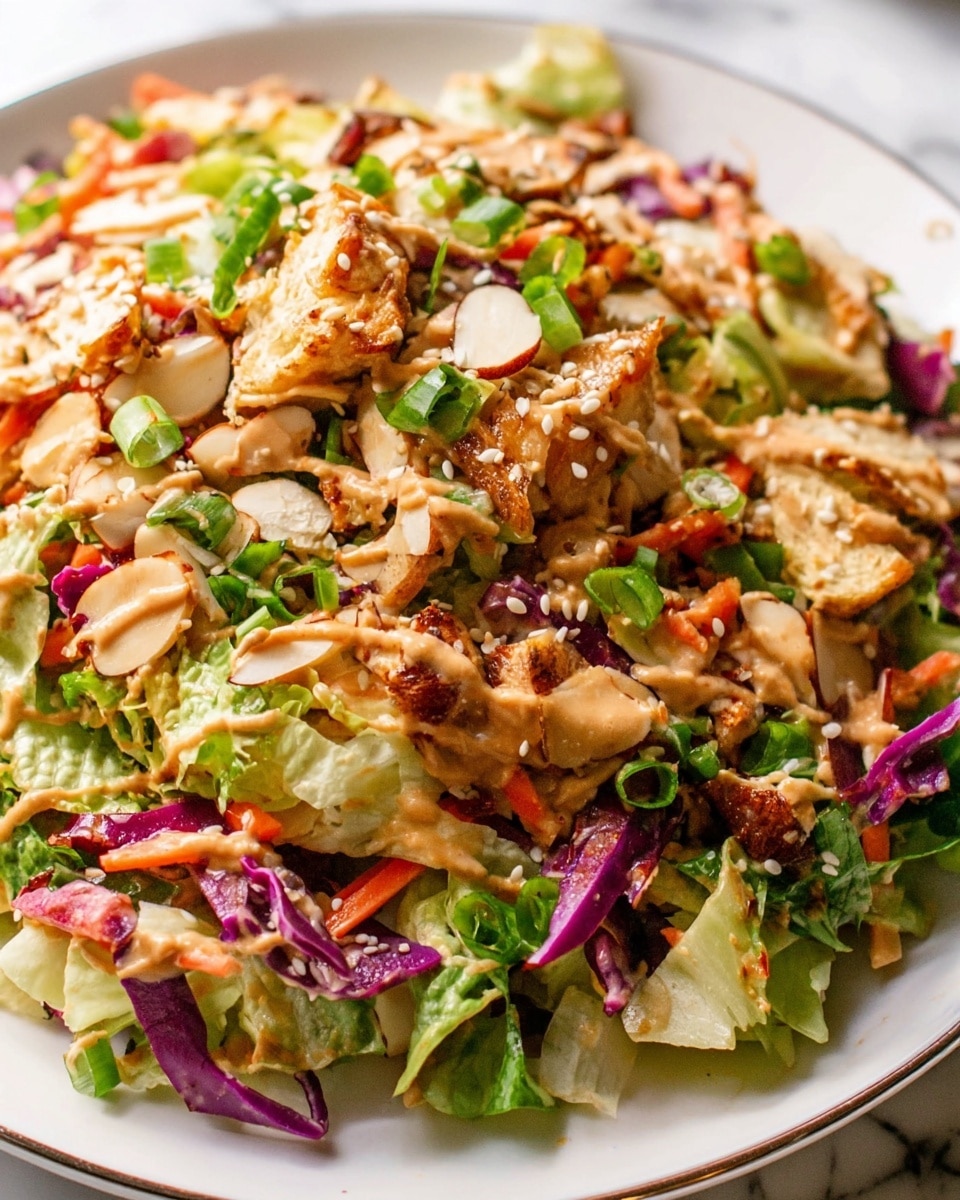 A close-up of a colorful salad served on a white plate with a thin silver rim, placed on a white marbled texture. The salad has multiple layers and textures: at the bottom, there are chopped green lettuce leaves and purple cabbage strips, mixed with thin orange carrot shreds; on top, there are browned chicken pieces, small green onion slices, and toasted almond slivers scattered over; a creamy light brown dressing is drizzled across the salad, along with beige sesame seeds sprinkled over all the layers. The salad looks fresh and vibrant, with a mix of crunchy and soft textures. Photo taken with an iphone --ar 4:5 --v 7