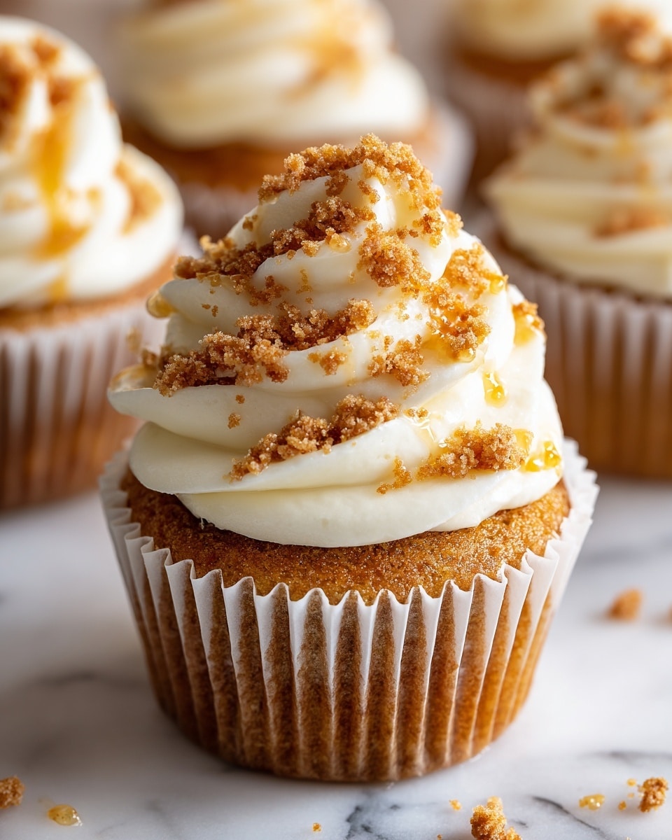 A close-up of a single cupcake with a golden-brown base wrapped in a white paper liner, topped with a thick swirl of smooth, creamy white frosting. The frosting is decorated with crunchy brown crumb-like bits sprinkled all over the top, with a light drizzle of golden syrup adding shine and texture. The cupcake sits on a white marbled surface with blurred cupcakes in the background. photo taken with an iphone --ar 4:5 --v 7