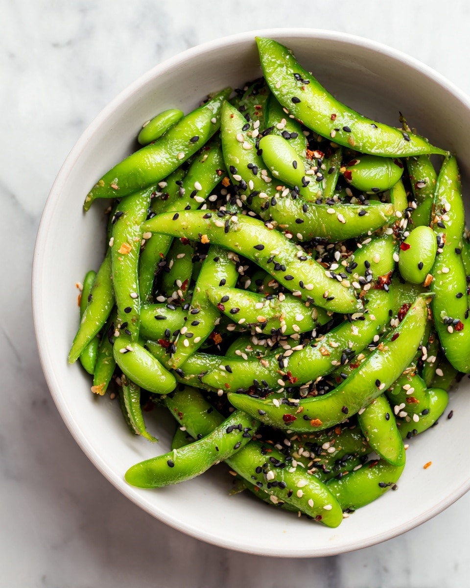 A white bowl filled with bright green edamame beans that are sliced lengthwise. The beans have a fresh, smooth texture and are sprinkled with black and white sesame seeds and some red chili flakes, adding small specks of contrast. The dish looks fresh and lightly seasoned, with a slight shine on the beans from a light dressing or oil. The bowl sits on a white marbled surface. photo taken with an iphone --ar 4:5 --v 7